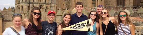Blind student in the middle holds a Nebraska Wesleyan pennant surrounded by other students in front of a castle.